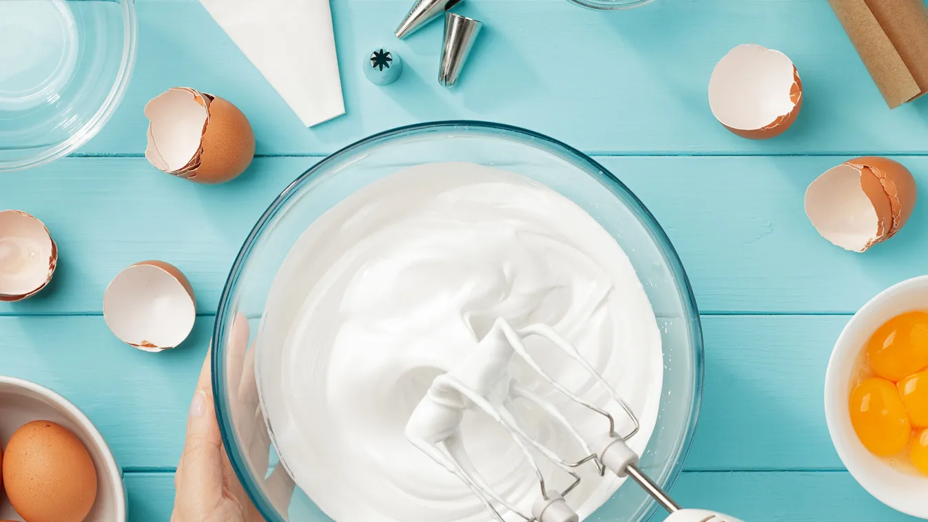Whipped egg whites cream to perfect peaks in glass bowl with mixer and hand on blue wooden table.