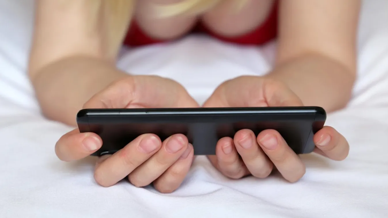 Blonde woman watching video on smartphone, sexy girl in negligee lying on the bed with mobile phone. Concept of porn, online addiction, sex dating