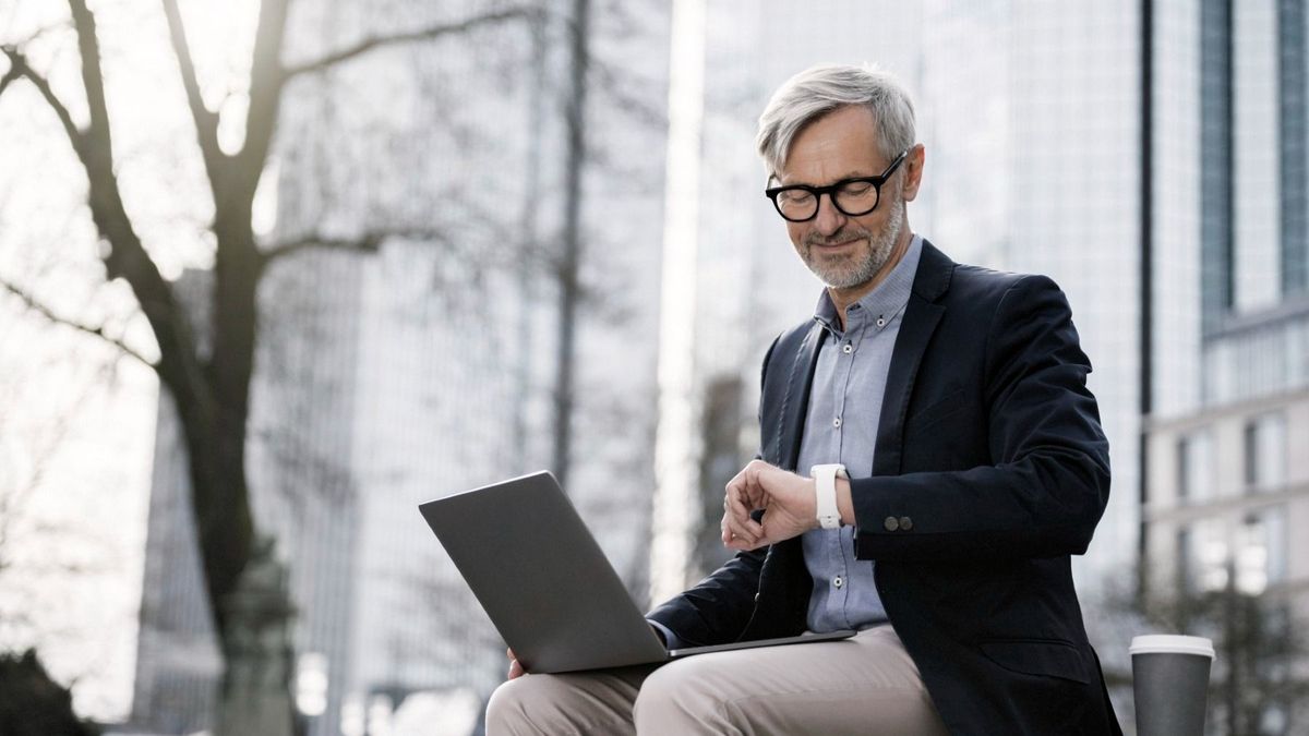 Grey-haired businessman working with laptop on wall in city looking at his smartwatch