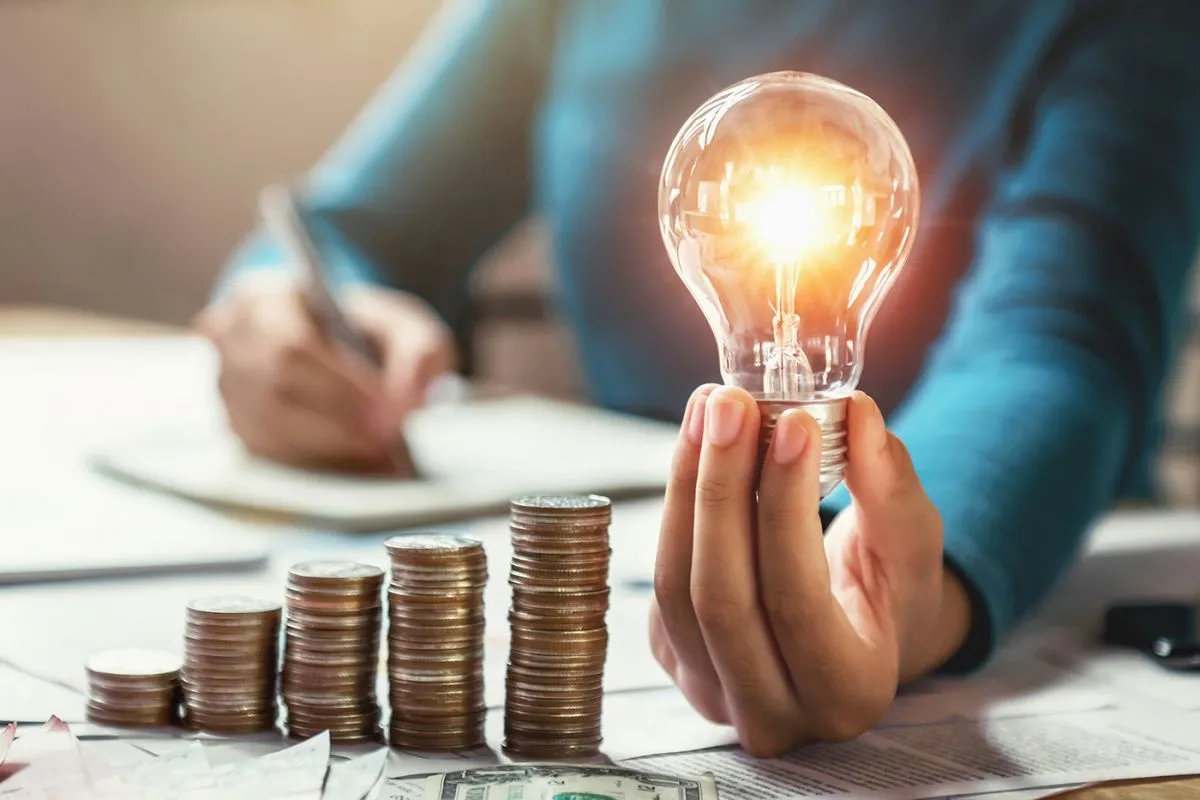 Business,Woman,Hand,Holding,Lightbulb,With,Coins,Stack,On,Desk.