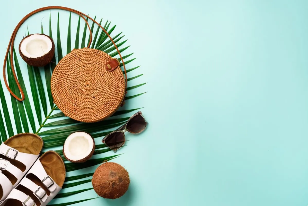 Stylish rattan bag, coconut, birkenstocks, palm branches, sunglasses on blue background. Banner. Top view with copy space. Trendy bamboo bag and white shoes. Summer fashion flat lay