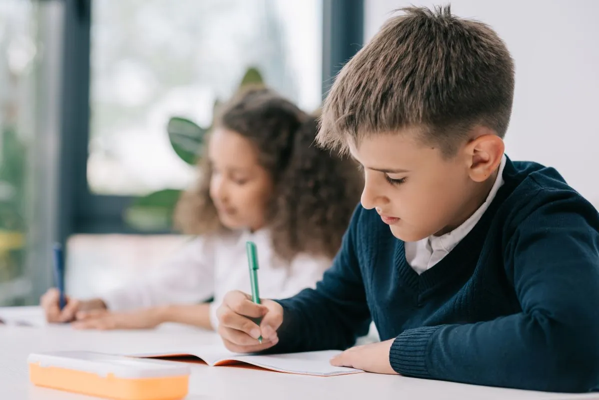 Concentrated,Schoolboy,Sitting,At,Desk,And,Writing,In,Exercise,Book