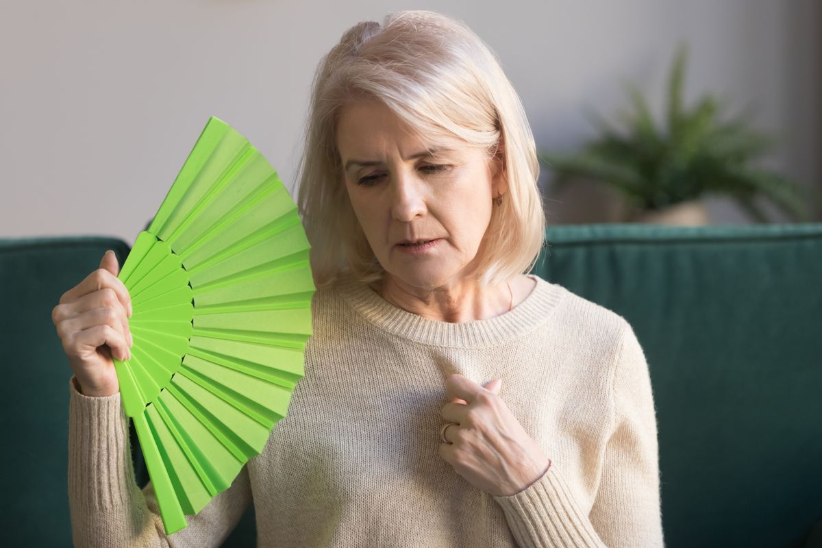 Overheated,Elderly,Woman,Sitting,On,Couch,Waving,Green,Fan,To
