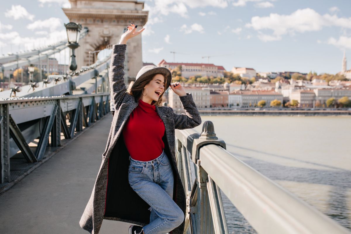Pretty young woman in blue jeans and long coat dancing on bridge near triumphal arch. Outdoor portrait of ecstatic brunette girl in red shirt travelling around Europe in autumn..