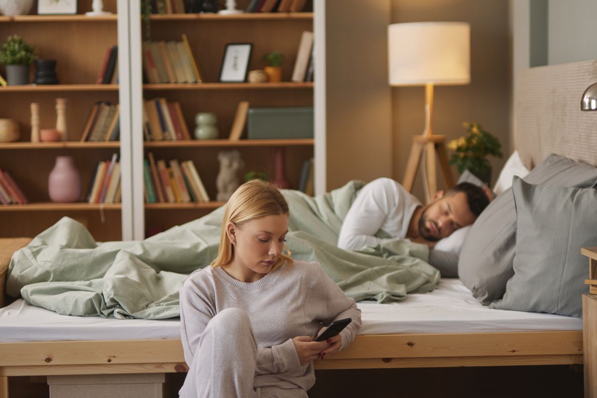 Woman,With,Smartphone,In,Bedroom,,Man,Sleeping,In,The,Background