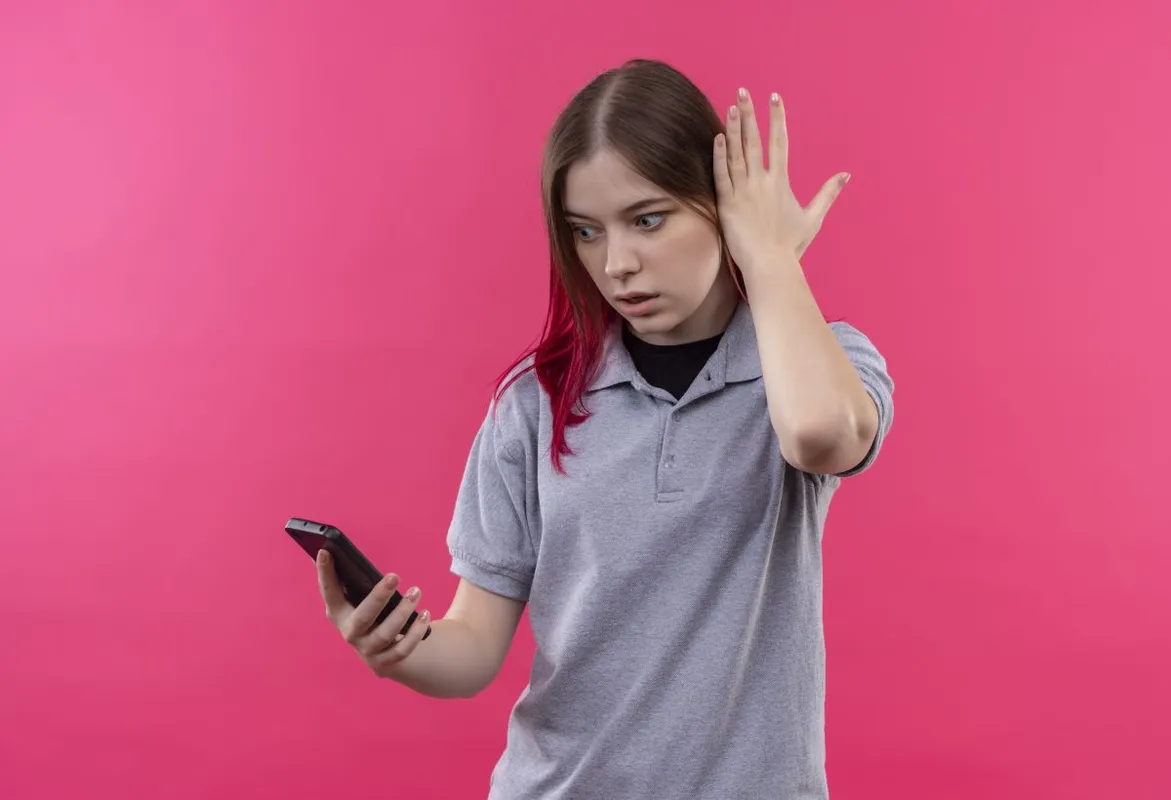 surprised young beautiful girl wearing gray t-shirt looking at phone in her hand on isolated pink background, android