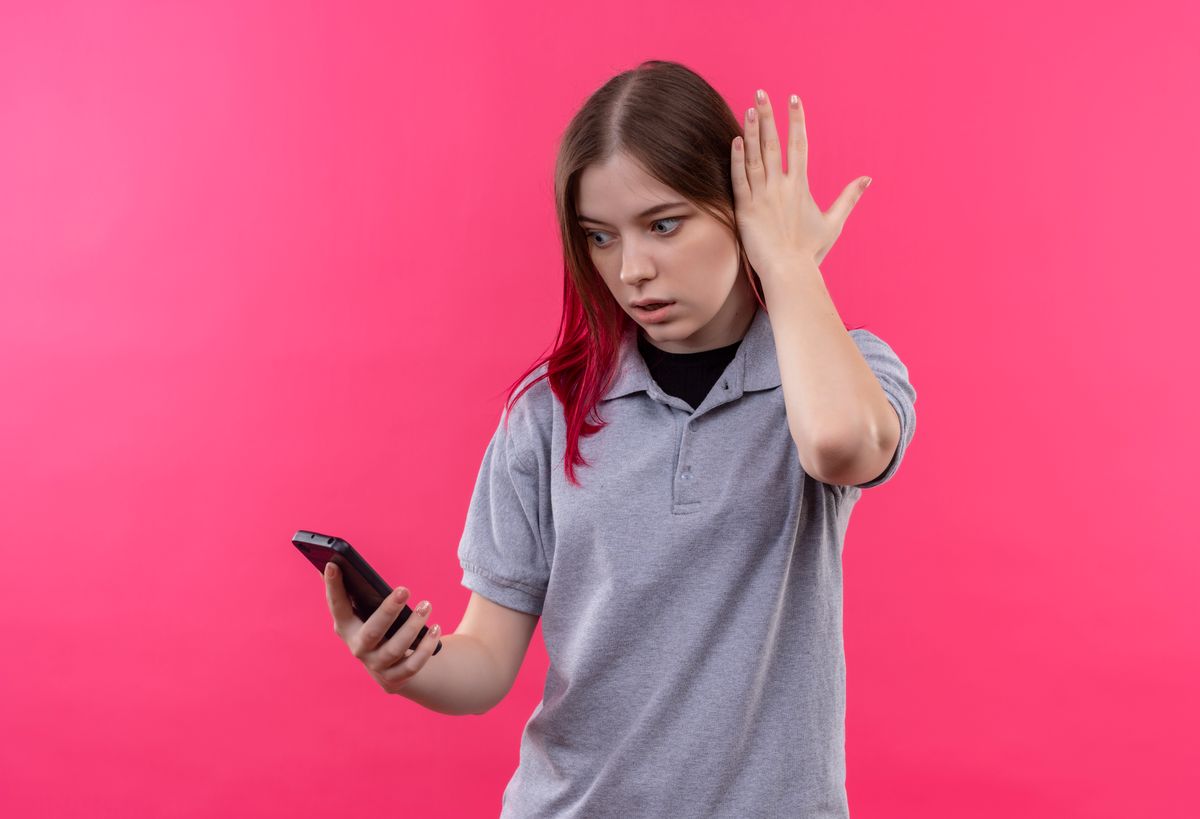 surprised young beautiful girl wearing gray t-shirt looking at phone in her hand on isolated pink background, android