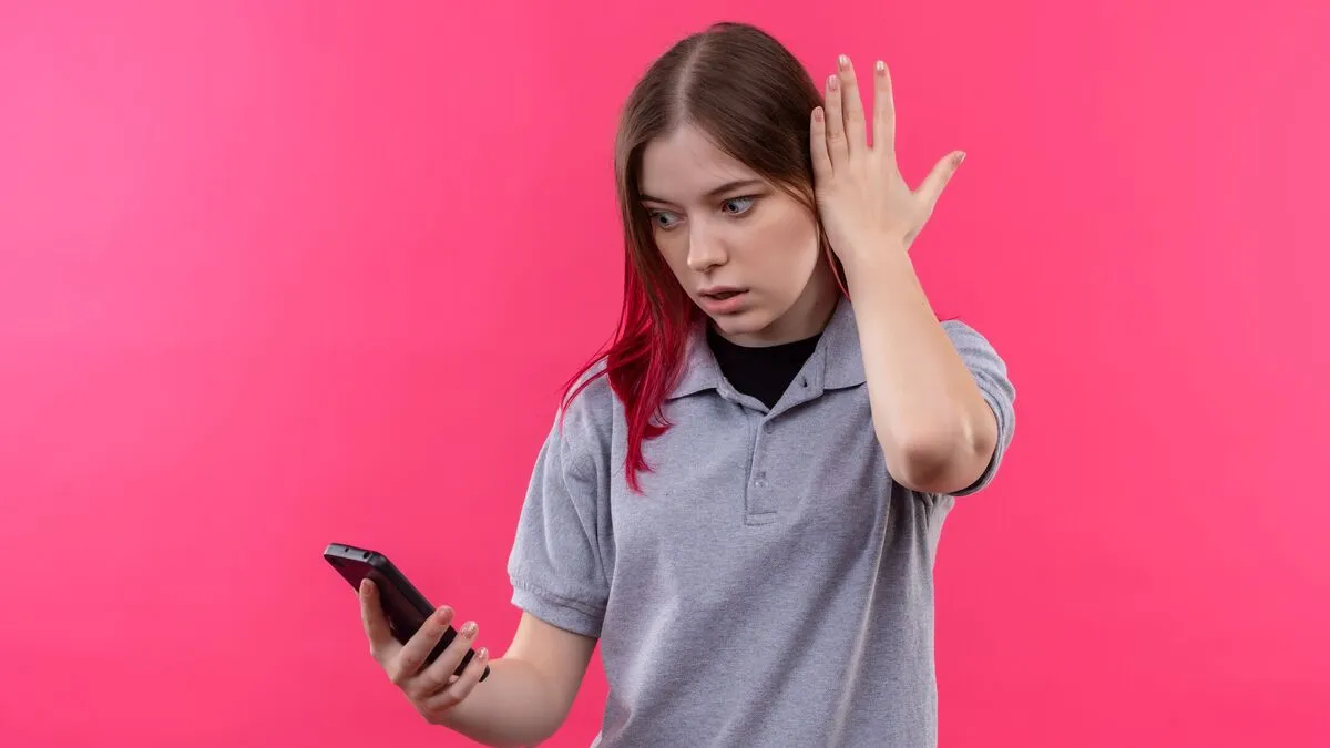 surprised young beautiful girl wearing gray t-shirt looking at phone in her hand on isolated pink background