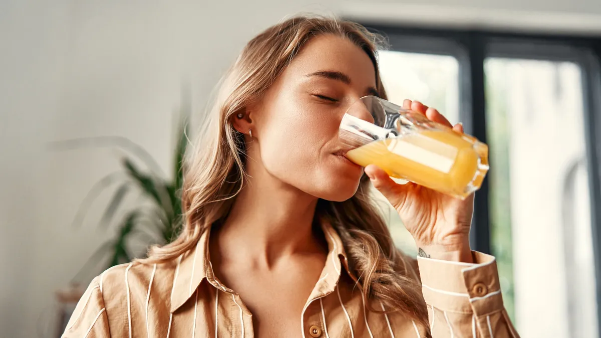 A,Young,Happy,Woman,Standing,In,The,Kitchen,Drinks,Orange
