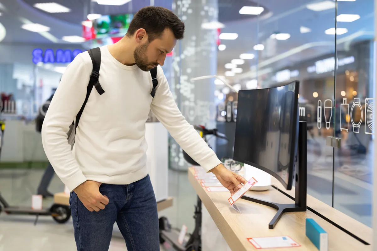 Minsk, Belarus - Nov 27, 2021: Photo of an adult handsome thinking brunet man with stubble in a white sweatshirt chooses a tv in an electronic store in a shopping mall. Shop concept